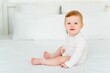 © Any Grant - A cute little smiling baby boy in a white bodysuit is sitting on a bed at home in a bright bedroom, a six-month-old baby with blue eyes is smiling or laughing
