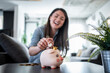© Migma_Agency - Happy woman saving money inserting coin in piggy bank at home