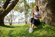 © matinee - Happy young woman in athletic wears sitting under a large tree, relaxing after a workout in the park.