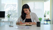 © wattana - Young businesswoman sitting at her desk, reading a financial document while taking notes in a planner.