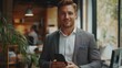 © liliyabatyrova - In a chic office setting, a well-dressed man holds a smartphone while flashing a confident smile, surrounded by people engaged in collaborative work and vibrant plants