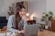 © Tj - Young Asian businesswoman working from home using laptop computer smiling and looking at screen sitting at a wooden table in her living room, working remotely or studying online
