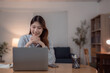 © Tj - Young Asian business woman working from home using laptop computer smiling and looking at screen, she is sitting at her desk in living room with warm light from lamp in the evening