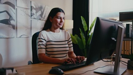 Brunette woman in striped shirt typing on keyboard and using mouse. Engaged in task. Manager finishing work after long day. Entering data into desktop. Dim sunlight shining through window.