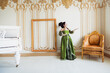 © deagreez - Elegant woman in green medieval-style dress in a luxurious baroque interior featuring a gold chair, empty frame, and white grand piano