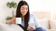 © Prostock-studio - Home Pastime. Smiling asian girl relaxing with smartphone and coffee on couch, browsing social media or messaging with friends, free space