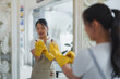© crizzystudio - Young woman in an apron putting on yellow rubber gloves, reflected in a mirror, getting ready to begin her cleaning shift and ensuring a hygienic environment for her clients