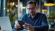 © Johannes - European smart businessman wearing glasses holding using cell mobile smartphone with laptop sitting at desk in office. Technology applications for business development growth and solutions concept.