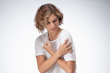 caucasian woman in a white shirt gripping her shoulder tightly with a painful expression, showing si