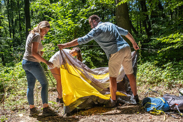 Fototapete - A family sets up a tent together in nature on a sunny summer day. They work as a team, helping each other to prepare their campsite.	
