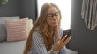 © Krakenimages.com - Young blonde woman with glasses using a smartphone in a cozy, modern living room.