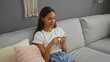 © Krakenimages.com - Young woman sitting on a gray couch in a living room, using a smartphone, and wearing casual clothes