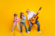 © deagreez - Cheerful grandfather playing guitar with two granddaughters singing on microphones against a lively yellow background.