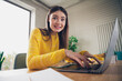 © deagreez - Young woman in a yellow sweater working on a laptop at home while smiling in a brightly lit interior workspace