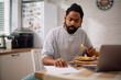 © Drazen - African American man analyzing his financial reports while having breakfast at home.