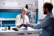 © DC Studio - Bearded man sits with elderly doctor in clinic office, reviewing health information on clipboard during routine checkup. Senior female physician checking medical history of male patient in hospital.