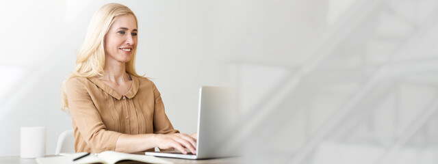 Aufkleber - A young woman with blonde hair sits at a clean workspace, typing on a laptop. She appears focused and engaged in her work, surrounded by a minimalist office environment with natural light, copy space
