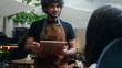 © Yuliia - Smiling Indian man Arabian latino guy friendly cafe worker barista in apron chef cafe customer service talk client order food lunch dinner in restaurant digital tablet serving visitor in coffee shop