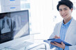 © Monet - A male doctor wearing a white lab coat stands in a modern clinic, smiling while holding a clipboard. On the computer screen behind him, chest X-ray images and medical data are displayed. The environme
