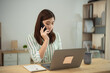 © WMSTUDIO - Asian Young woman engaged in a phone conversation while working on a laptop at a stylish home office with a modern aesthetic and minimalistic decor