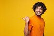 © ViDi Studio - Side view young smiling Indian man wear orange t-shirt casual clothes point thumb finger back aside on blank empty area mockup isolated on plain yellow background studio portrait. Lifestyle concept.