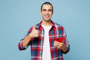 Young happy middle eastern man he wear red shirt casual clothes hold in hand use mobile cell phone show thumb up isolated on plain pastel light blue cyan background studio portrait. Lifestyle concept.