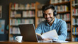 © GG Kenji - Flat Smiling young man with wireless headset studies e learning on laptop in library taking notes watching webinar online course video call concept as Young man in library with wireless headset studyi