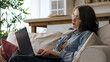 © yavdat - Seated on a sofa, a woman in casual attire with glasses writes in a notebook, her gaze fixed on a laptop screen. This photo emphasizes the dedication and focus required for successful remote work or