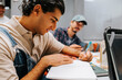 © Maskot - Smiling young man writing on diary while sitting in classroom at college