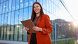 © PeopleVideos - Smiling businesswoman holding clipboard standing in front of modern office building at sunset