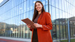© PeopleVideos - Smiling businesswoman holding clipboard standing in front of modern office building