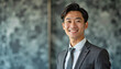 © f_bossa - A portrait headshot photo of a friendly professional CEO executive business worker: A smiling Asian man in a suit and tie confidently poses against a textured gray wall, projecting a professional