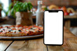 © Stas - Smartphone with blank white screen displayed on a wooden table next to a pizza for showcasing food delivery apps, concept: food, phone, pizza, delivery, app