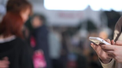 Woman is holding a cell phone in her hand. She is looking at the screen, possibly texting or browsing the internet. The scene takes place in a public area, with other people around her