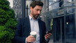 © Yuliia - Male manager in city near office drinking coffee looking away relaxed thinking dreaming smiling calm look check time with mobile phone Caucasian man business guy employer businessman run late to work