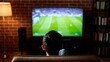 © Justice - a person sitting on a couch in a cozy living room, watching a soccer match on a large flat-screen TV. The room has a brick wall and a bookshelf, creating a comfortable atmosphere for watching TV.