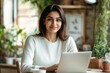 © yang - Indian woman sits at table with laptop engaging in professional tasks, working in homeoffice, feel happy and satisfied with productive workflow, take break, look at camera, posing. Freelancer portrait