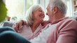 © Curioso.Photography - Happy senior couple sits close together on a couch, smiling warmly at each other in a cozy home setting.