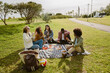 © Drobot Dean - Group of five adults sitting on a picnic blanket in a park during daytime, eating fruit and snacks. A Black man with an afro wears a green jacket, a white man with curly hair wears a cream jacket.