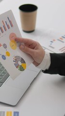 Poster - In a stylish office, a dedicated businesswoman reviews printed charts and graphs, deeply engaged with data on her laptop, highlighting her analytical approach to meetings and strategic planning