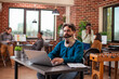 © DC Studio - Professional male analyst reviewing research data on device and managing work tasks while recovering from an injury. Bearded man with a neck brace using laptop in brick wall workspace.