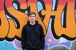 © velikiyzayats - A teenage male standing confidently in front of vibrant graffiti art on a wall, showcasing a mix of colors and styles, highlighting urban street culture.