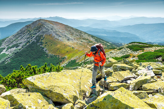 Bearded backpacker in an orange jacket trekking over large boulders on a rugged mountain ridge. Panoramic mountain views in the background.