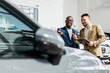 © Vladislav - A car dealer in a modern car dealership advises an African-American customer on buying or leasing a new car. The concept of an expensive purchase or lease of a modern car.