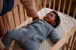 © Seventyfour - High angle shot of Black mother putting sleeping baby girl dressed in knitted jumpsuit into crib while taking care of child in home setting