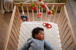 © Seventyfour - Cropped shot of little Black girl dressed in blue knitted jumpsuit sleeping on starry blanket in baby crib with musical mobiles, hanging above child in home setting