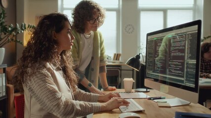 Two female IT specialists collaborating on software development project, discussing code displayed on desktop computer monitor in modern office