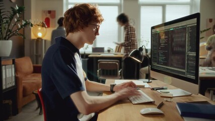 Young male developer typing on computer keyboard and examining code on desktop monitor, working in modern open plan office with colleagues moving in background. Time lapse video