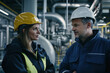 © Lady O - Two engineers talking to each other, wearing working clothing and protective helmets, in a modern factory full of pipes and machines.