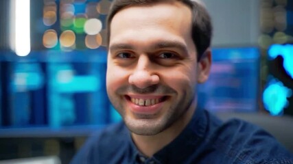 Cheerful young man smiling confidently in tech office with data screens in background. Perfect for themes like IT professional, software engineer, success, or startup culture.